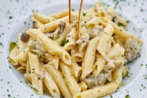  A bowl of penne pasta topped with sautéed mushrooms and melted cheese, garnished with herbs.